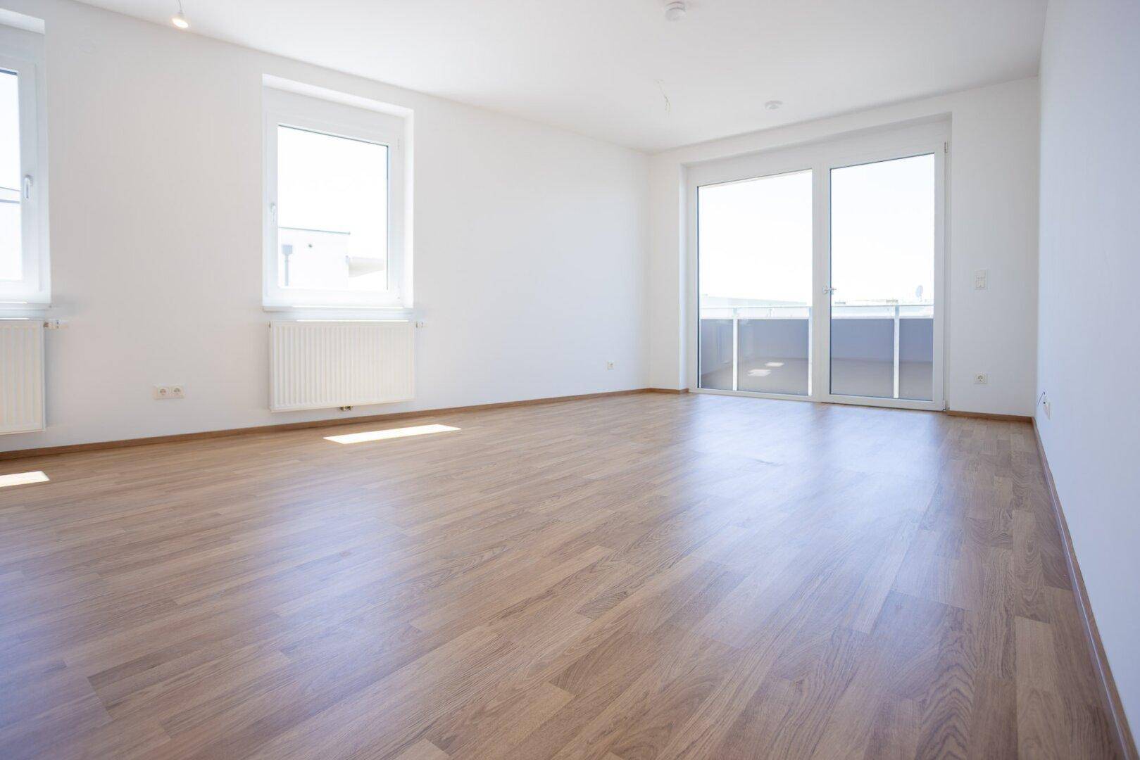 Heller Wohnraum mit Laminatboden, weißen Wänden und großem Fenster mit Zugang zum Balkon.
