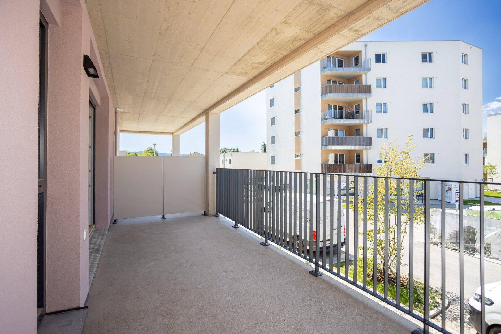 Langer Balkon mit Sichtschutz und Ausblick auf die moderne Wohnanlage und den Himmel.