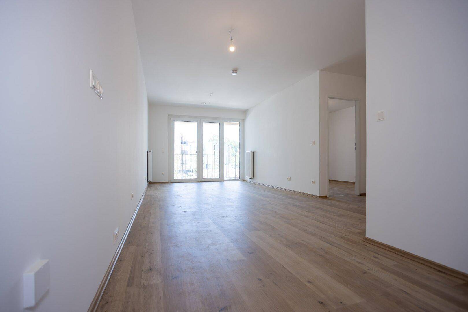 Weitläufiges, unmöbliertes Zimmer mit hellem Laminatboden und Balkonzugang in einem Neubau.