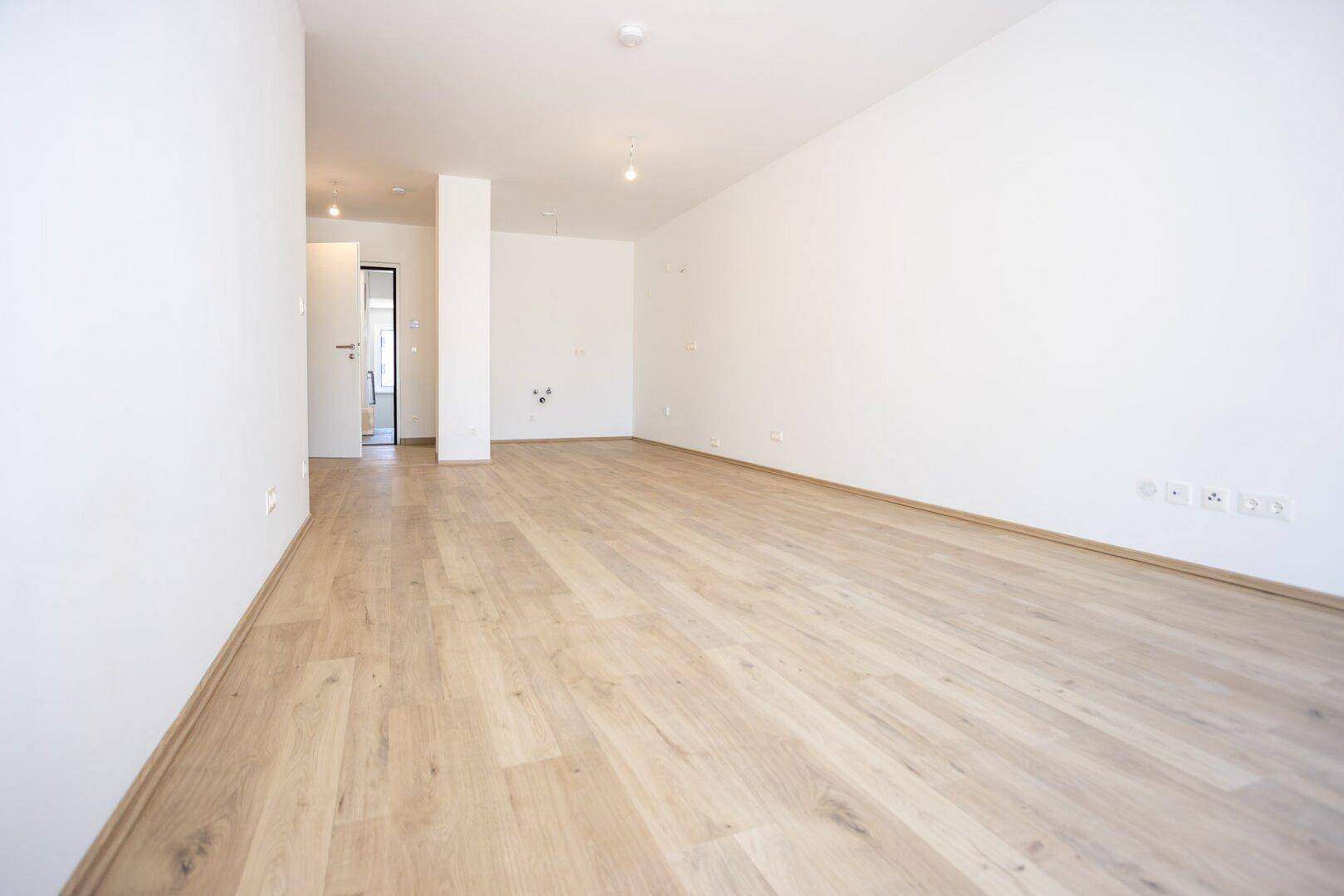 Geräumiges, unmöbliertes Zimmer mit hellem Laminatboden und weißen Wänden in einem Neubau.