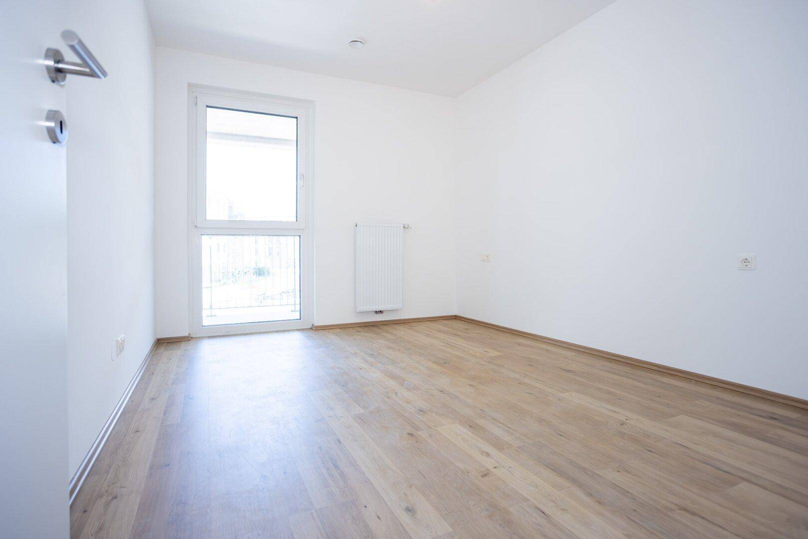 Helles Schlafzimmer mit Laminatboden und Fenster mit französischem Balkon in einem Neubau.
