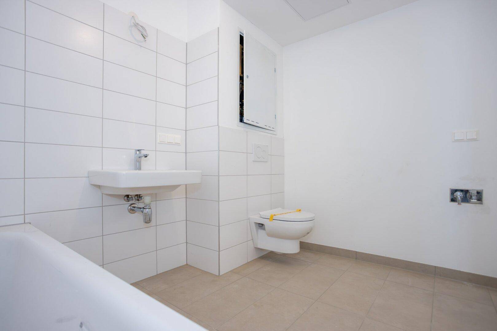 Helles Badezimmer mit weißen Fliesen, Waschbecken, wandhängendem WC und Badewanne, bereit zur Nutzung.