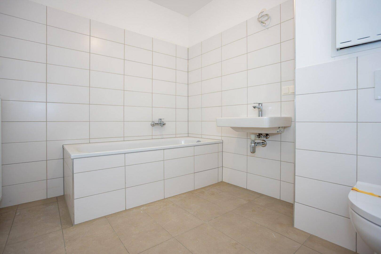 Sauberes Badezimmer mit Badewanne, kleinem Waschbecken und weißen Wandfliesen, funktional und hell.