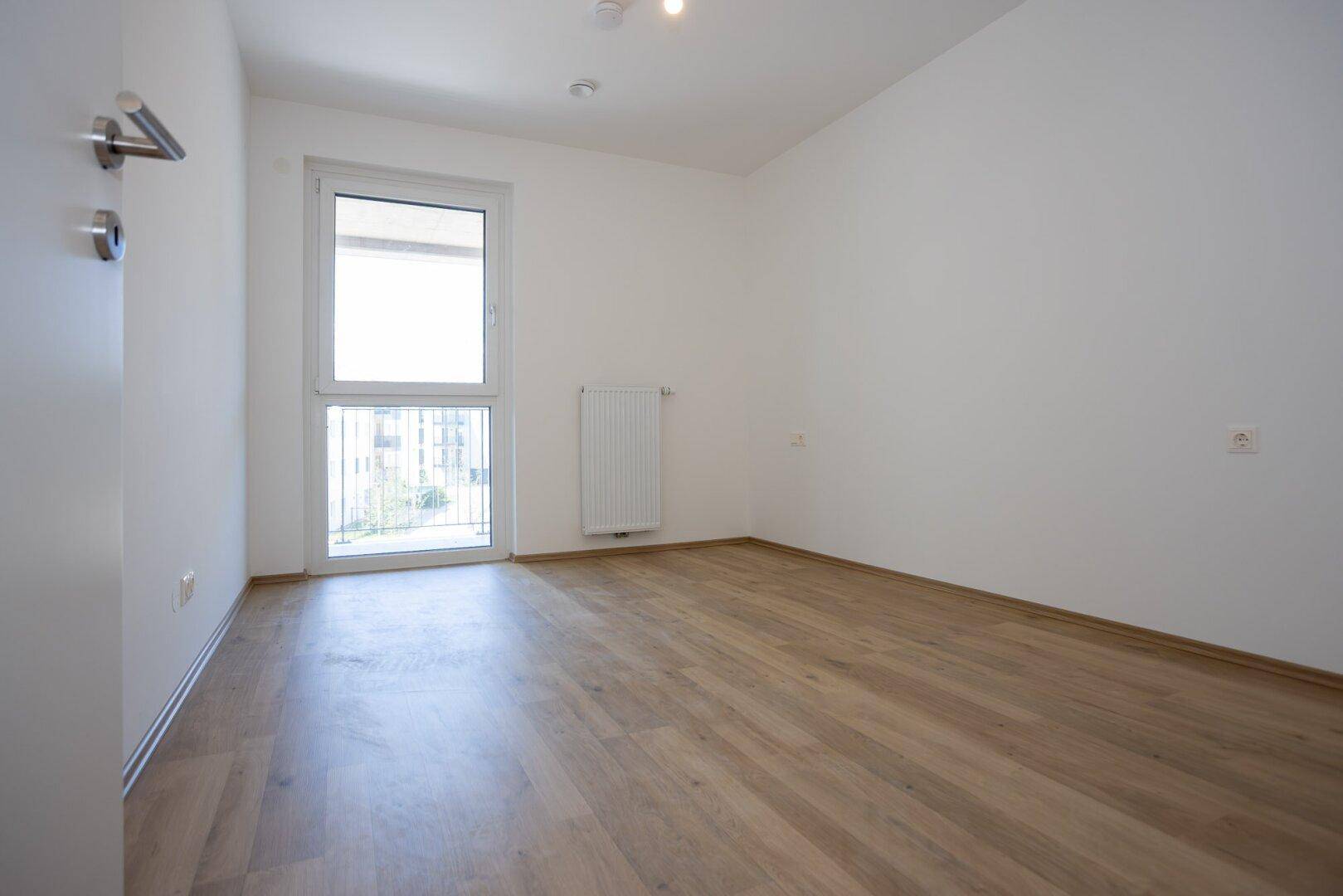 Helles Schlafzimmer mit Laminatboden, Heizkörper und französischem Balkon, ideal für ein Kinderzimmer oder Büro.