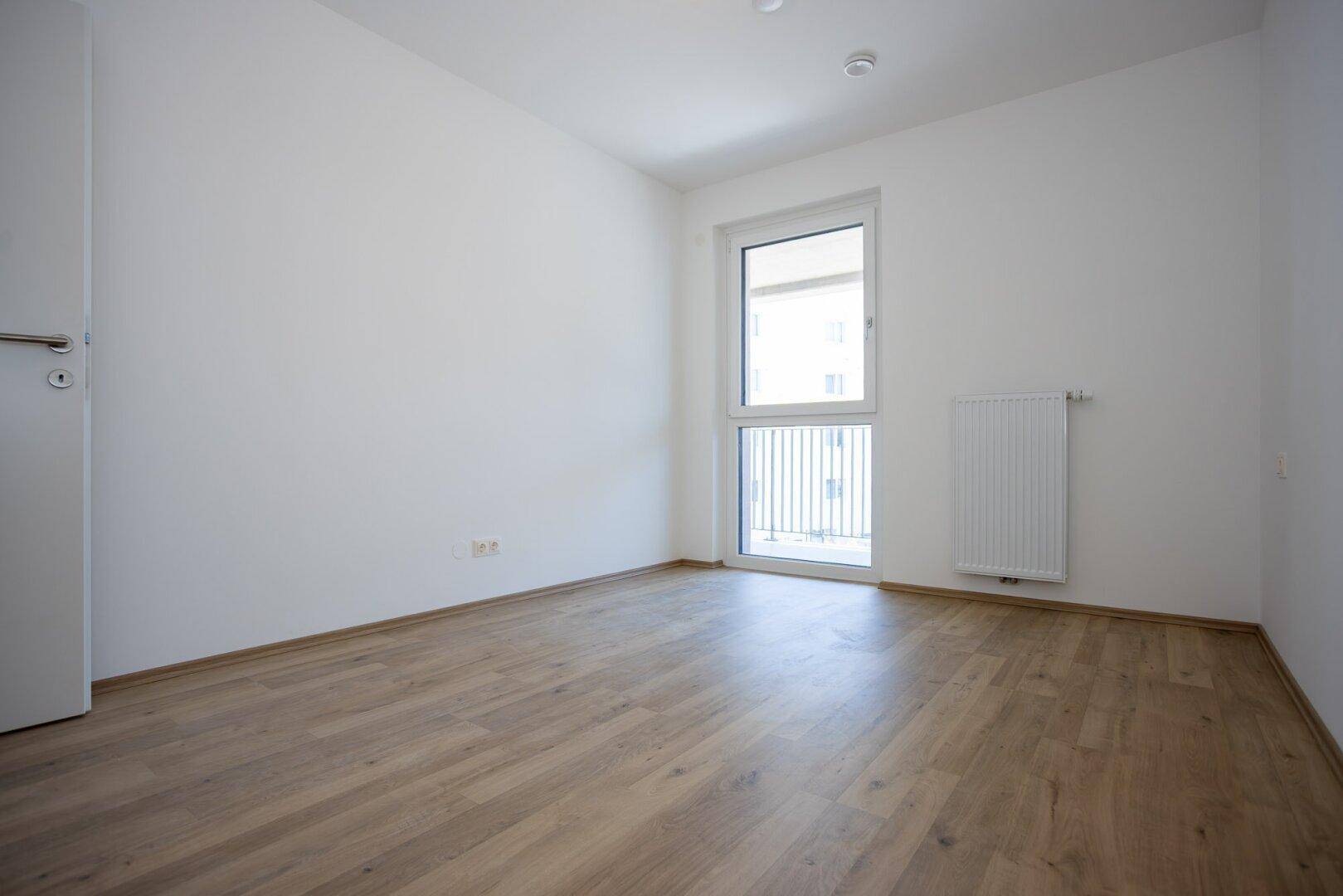 Unmöbliertes Schlafzimmer mit Laminatboden, weiß gestrichenen Wänden und Fenster mit französischem Balkon.