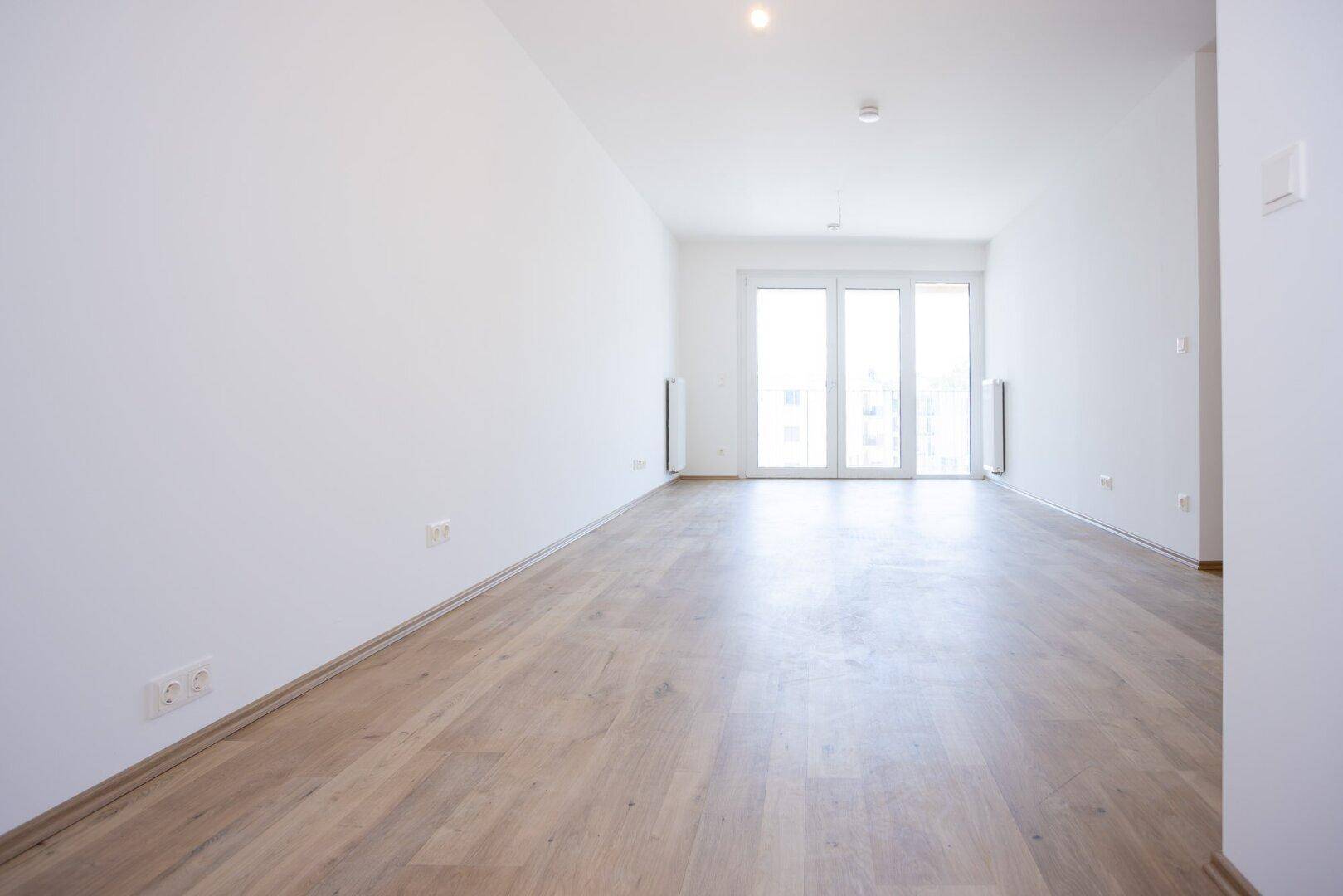 Lichtdurchfluteter Wohnraum mit großen Fenstern und hellem Laminatboden in einem Neubau.