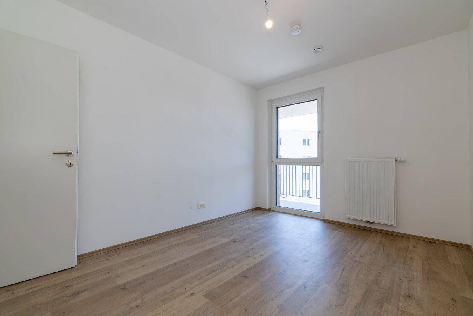 Unmöbliertes Schlafzimmer mit hellem Laminatboden und Fenster mit Blick nach draußen.