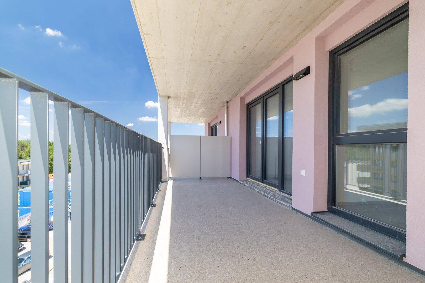 Langer Balkon mit Betonboden und moderner grauer Brüstung, bietet einen sonnigen Außenbereich.