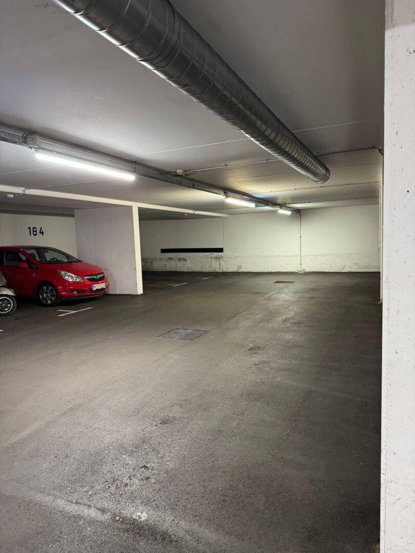 Innenansicht einer Tiefgarage mit einem geparkten roten Auto, Betonboden und sichtbaren Lüftungsrohren.
