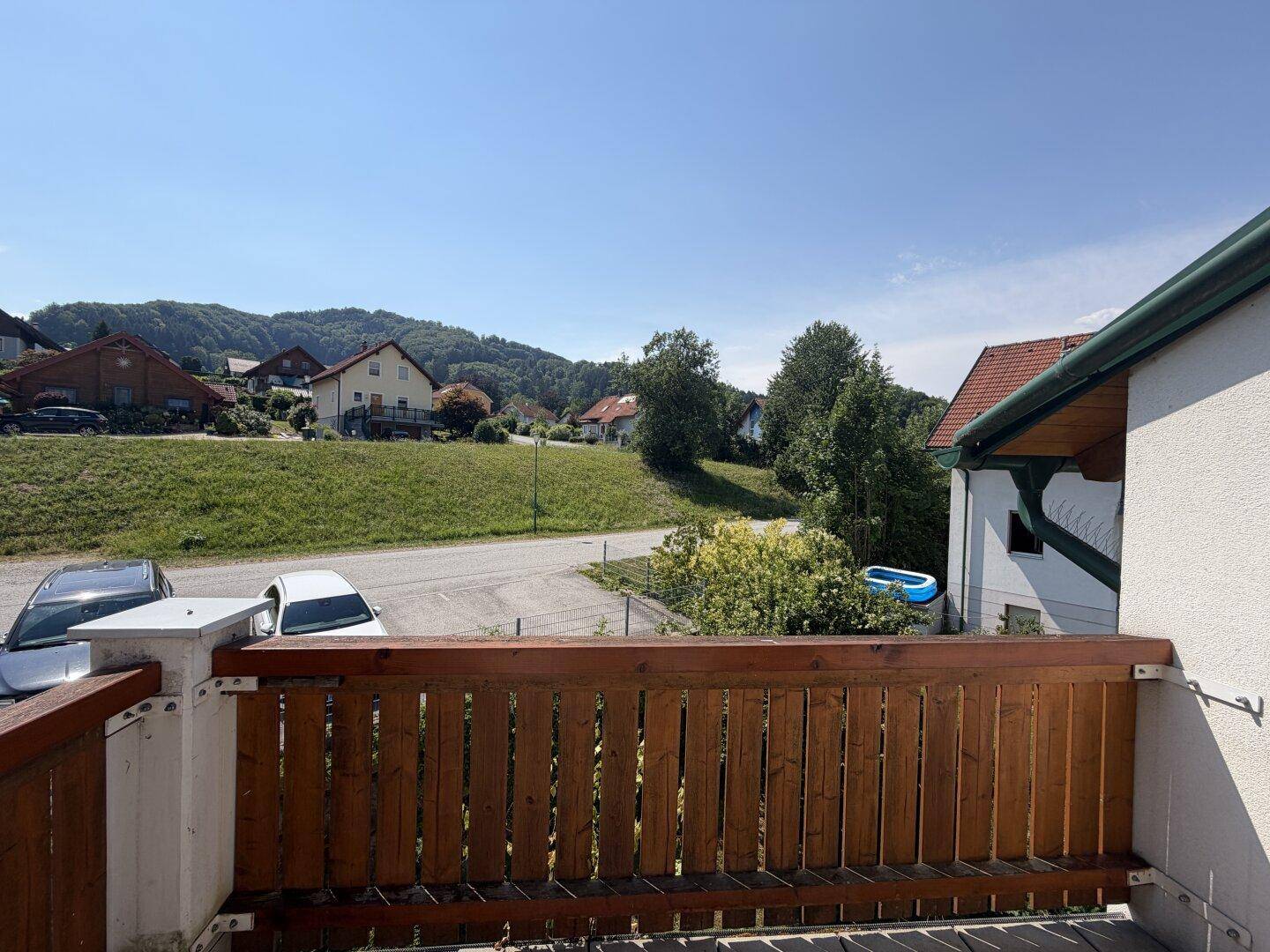 Sonniger Balkon mit Holzgeländer und weitem Blick über die grüne Umgebung und Nachbarhäuser.