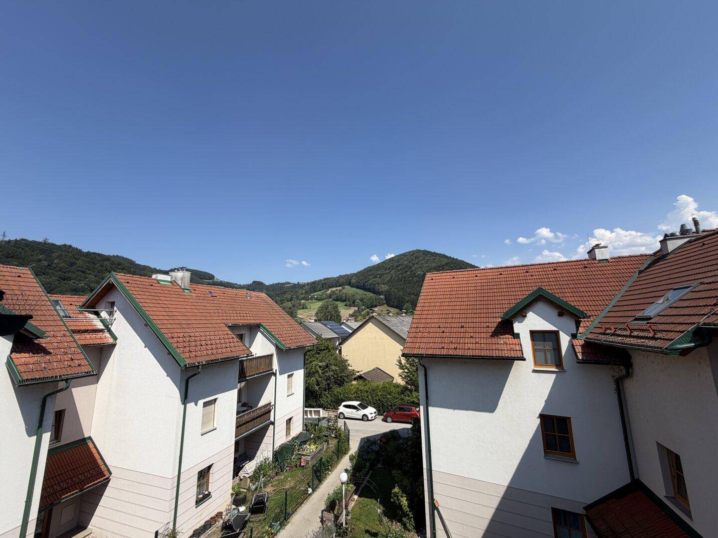 Ansicht mehrerer Wohnhäuser mit roten Ziegeldächern und grüner Umgebung vor einer Hügellandschaft.