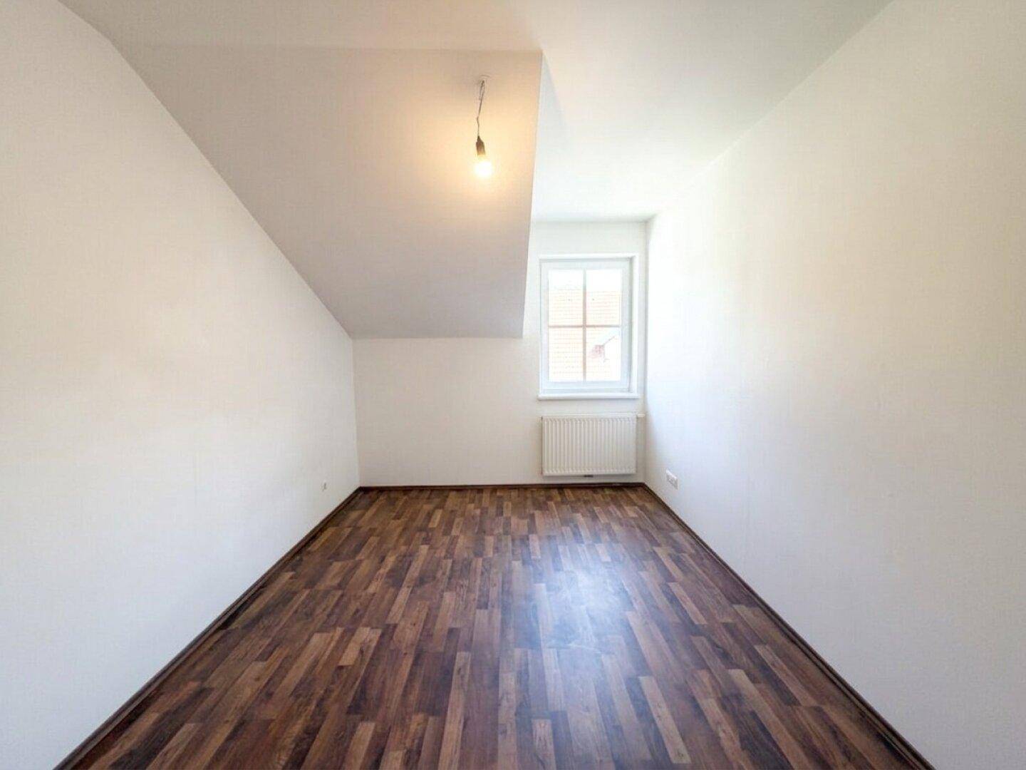 Gemütliches Dachzimmer mit dunklem Laminatboden, weißen Wänden und einem Fenster in der Schräge.