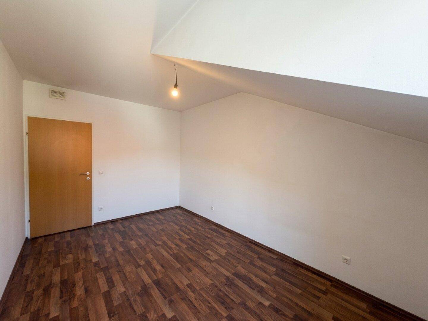 Dachzimmer mit dunklem Laminatboden, weißen Wänden und einer Holztür, ideal als Schlafzimmer.