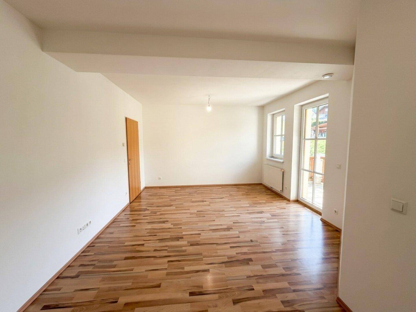 Geräumiges, unmöbliertes Zimmer mit hellem Laminatboden und viel Tageslicht durch Fenster und Tür.