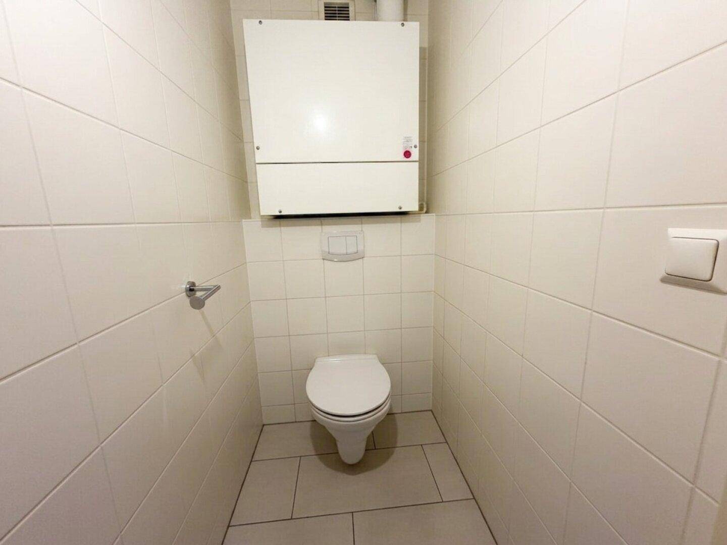 Separates WC mit weißen Fliesen, wandhängender Toilette und Belüftungssystem.