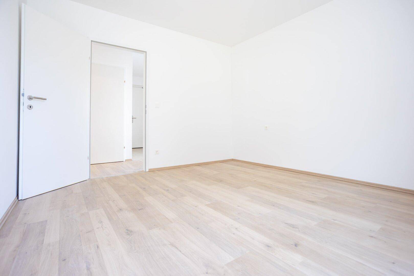 Leeres Zimmer mit hellem Laminatboden und weiß gestrichenen Wänden, bereit zur individuellen Gestaltung.