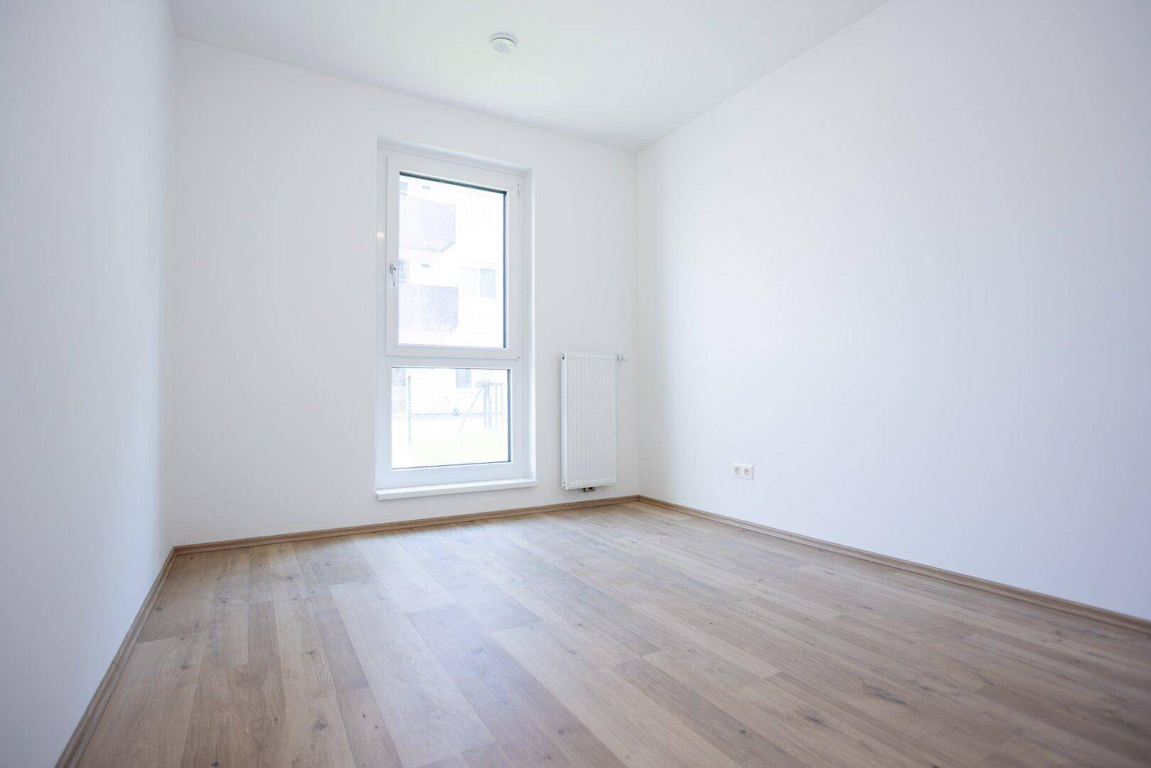 Freundliches Zimmer mit hellem Laminatboden, weiß gestrichenen Wänden und einem hohen Fenster.