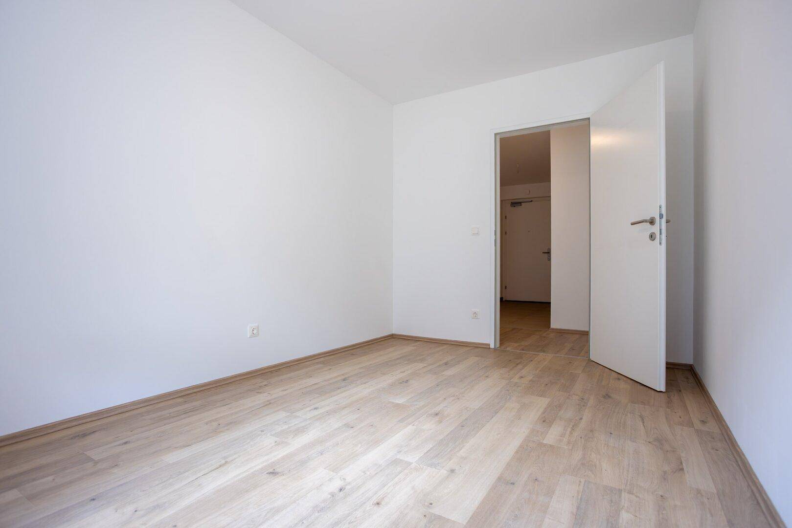 Schlichtes, leeres Zimmer mit Laminatboden und weiß gestrichenen Wänden, mit Zugang zum Flur.