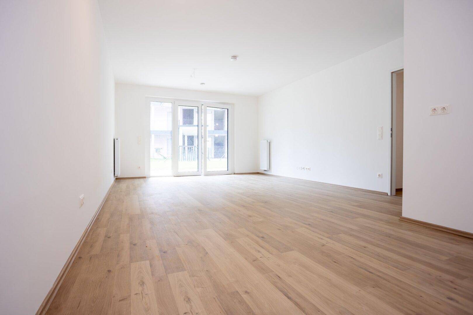 Helles, leeres Wohnzimmer mit modernem Laminatboden und Zugang zu einem Außenbereich.