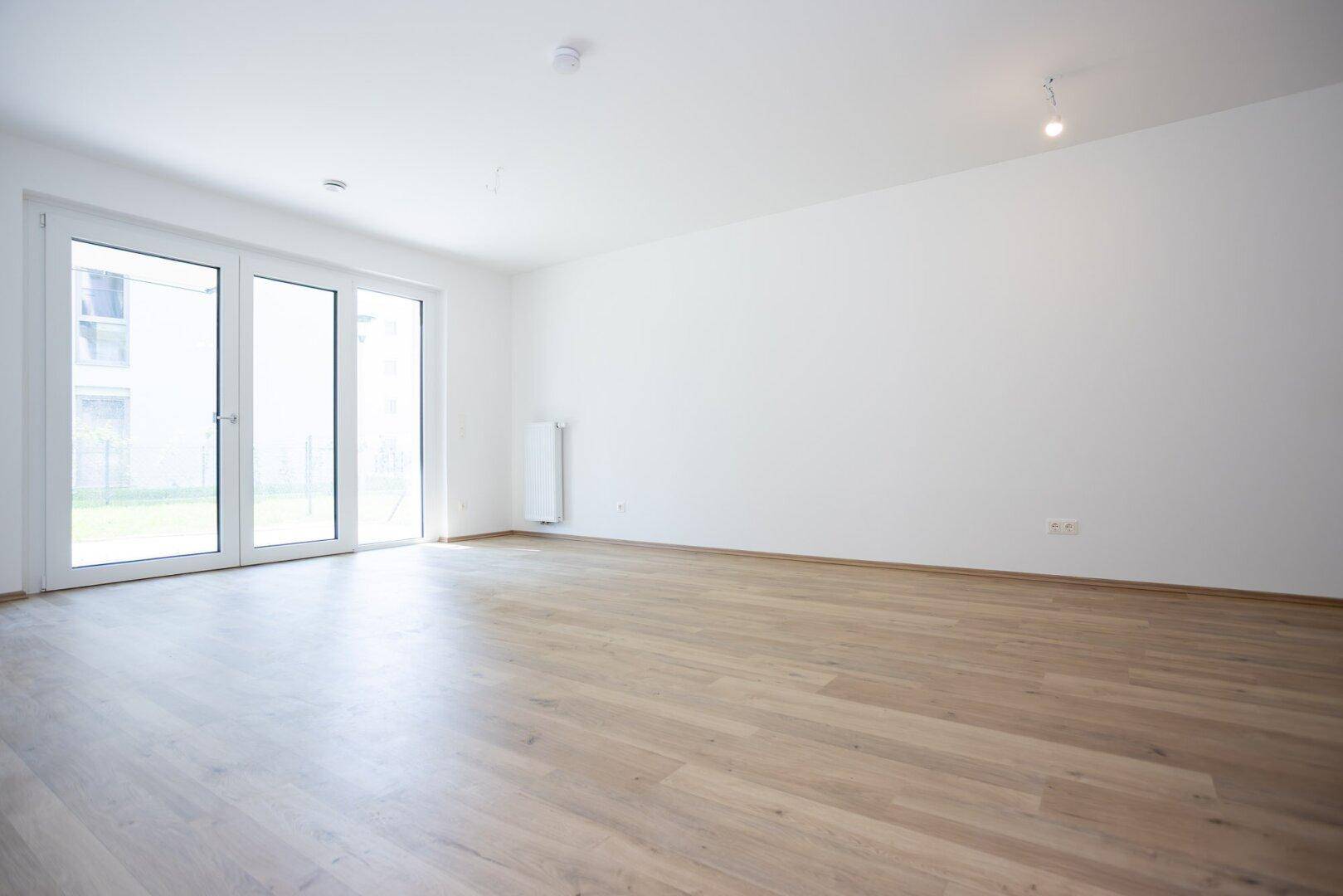 Heller, leerer Wohnraum mit großen Terrassentüren und hellem Holzboden.