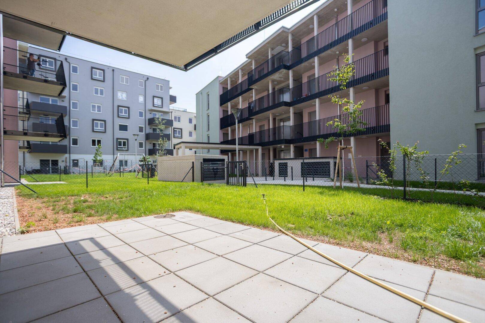 Gemeinschaftlicher Gartenbereich mit gepflasterter Terrasse und modernen Wohngebäuden im Hintergrund.