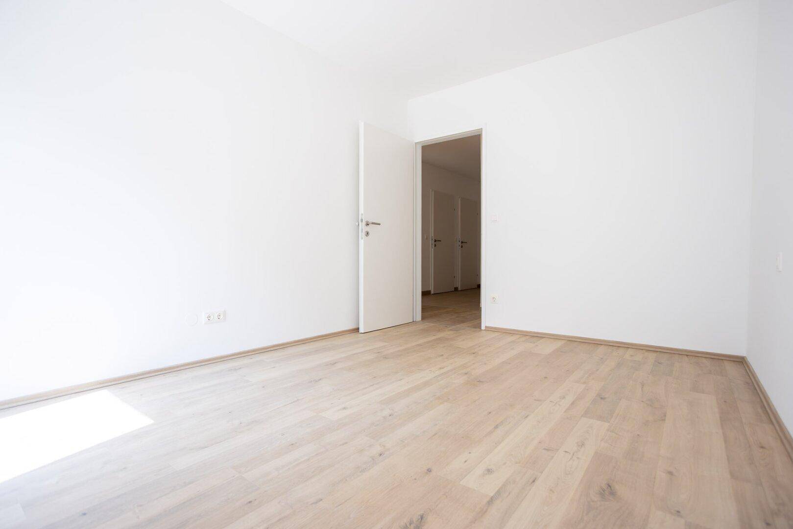 Leeres Zimmer mit offener Tür, hellem Holzboden und weißen Wänden.