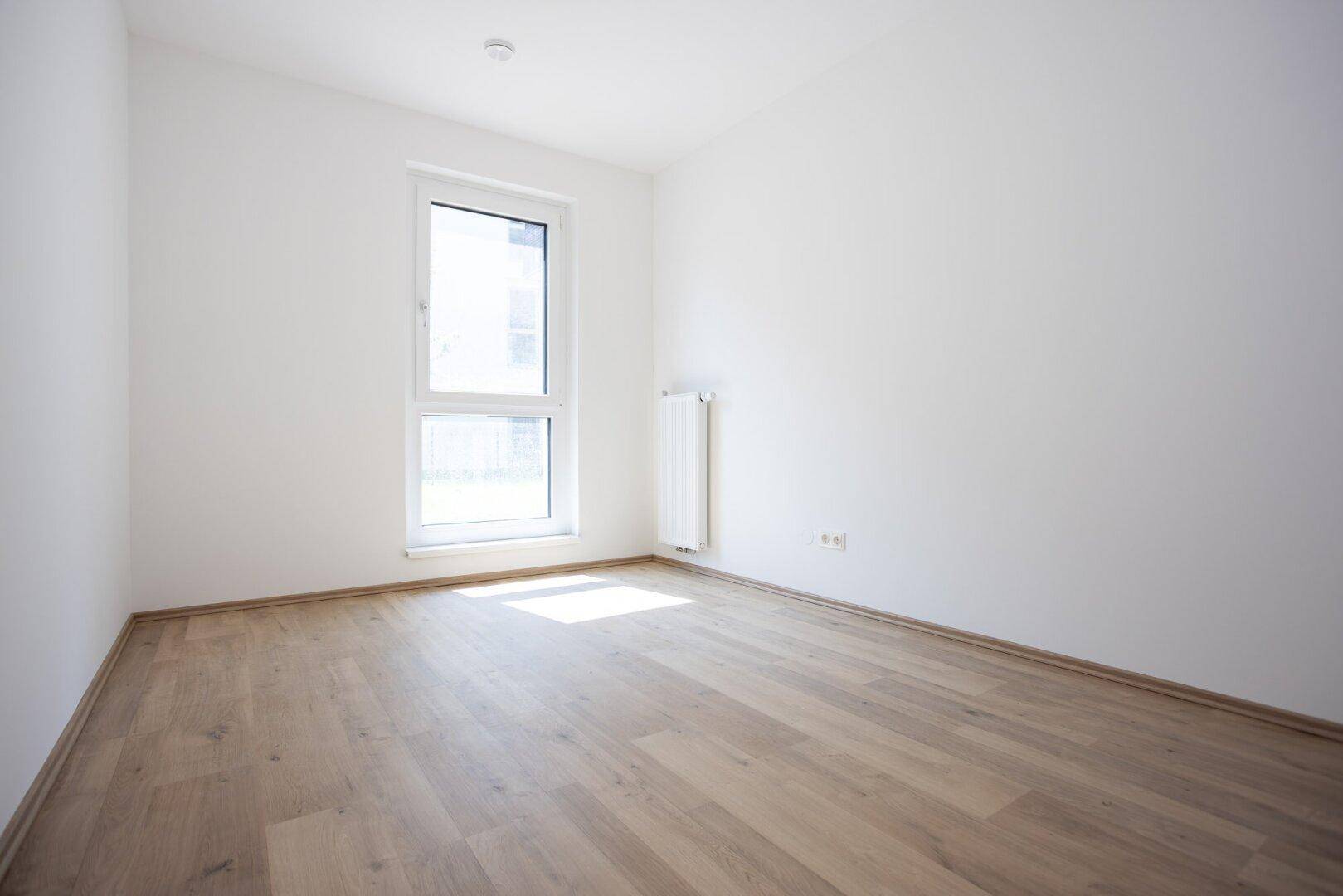 Helles Zimmer mit großem Fenster, hellem Holzboden und weiß gestrichenen Wänden.