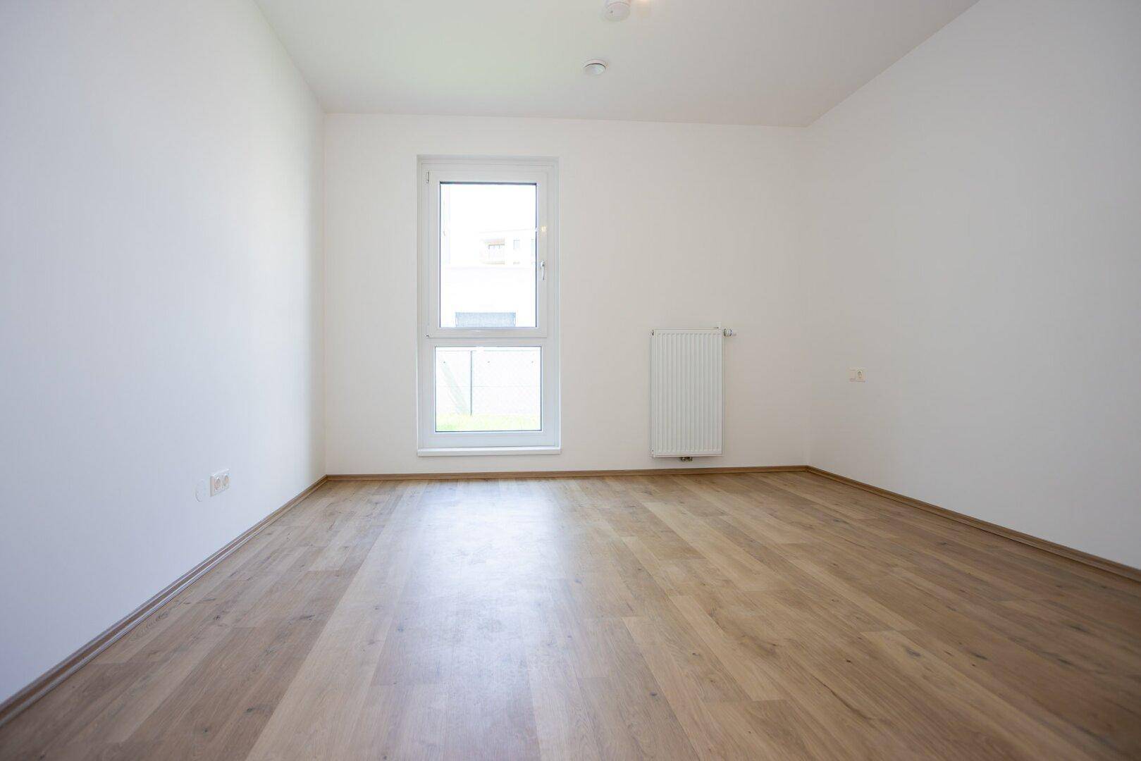 Helles, unmöbliertes Zimmer mit Laminatboden und einem Fenster mit Blick ins Grüne.