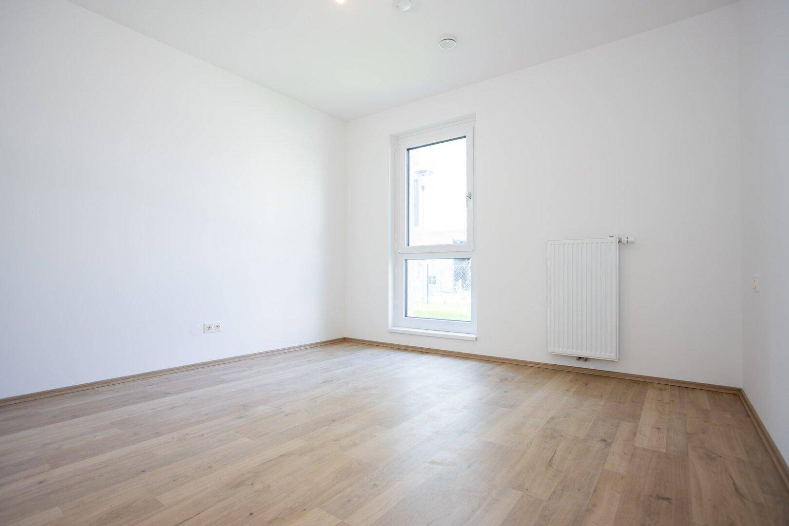 Leeres Zimmer mit hellem Laminatboden und einem Fenster, das viel Tageslicht hereinlässt.