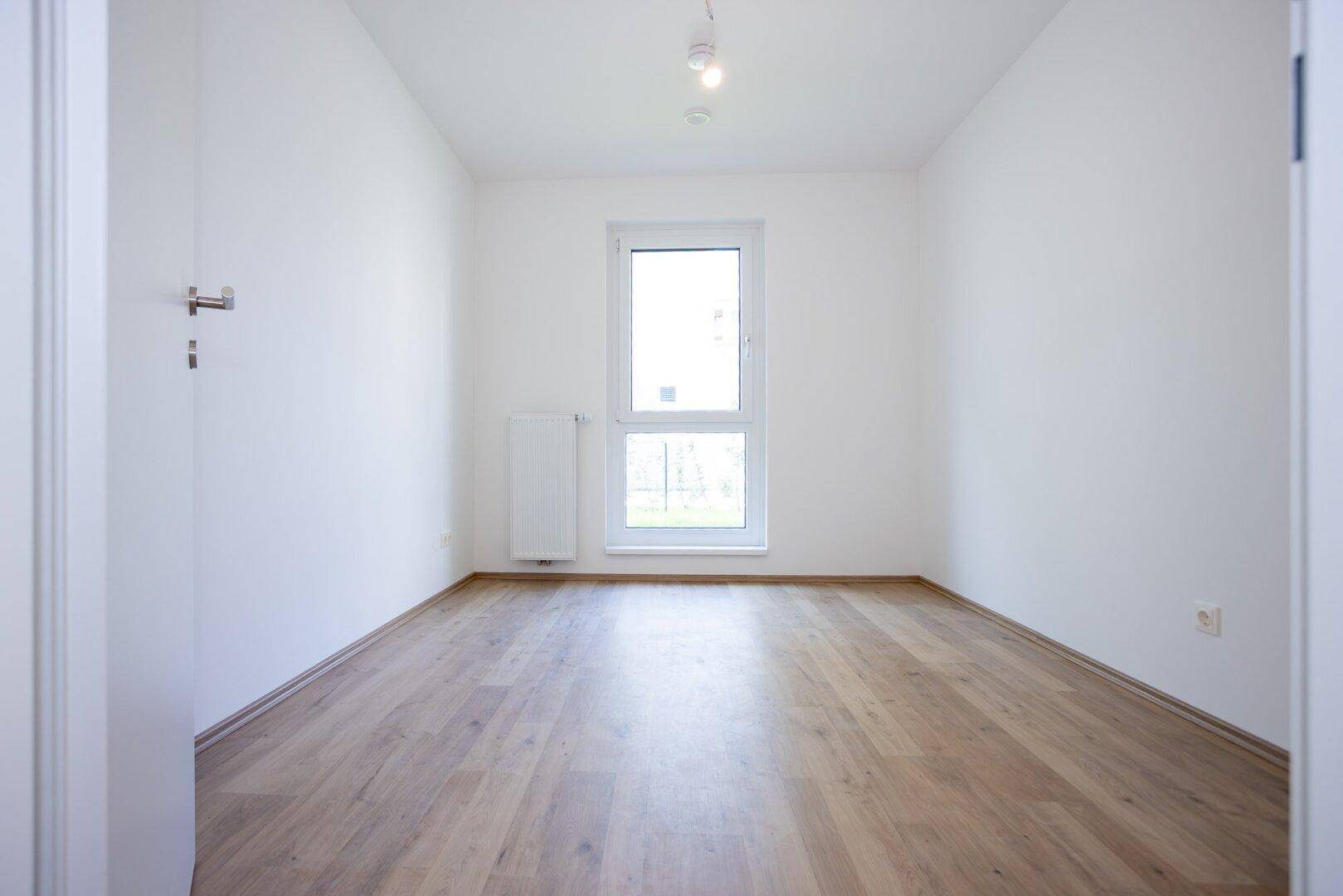 Helles, leeres Schlafzimmer mit Laminatboden und einem großen Fenster in einem Neubau.