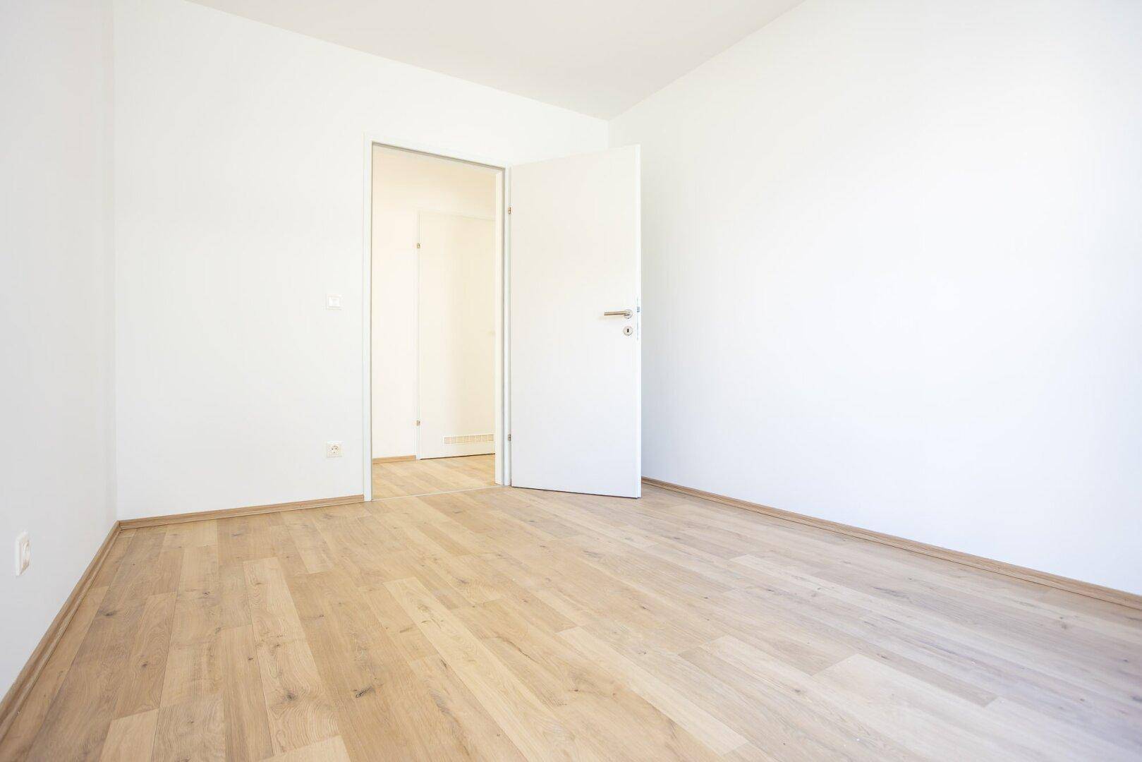 Helles, unmöbliertes Zimmer mit Laminatboden und weiß gestrichenen Wänden.