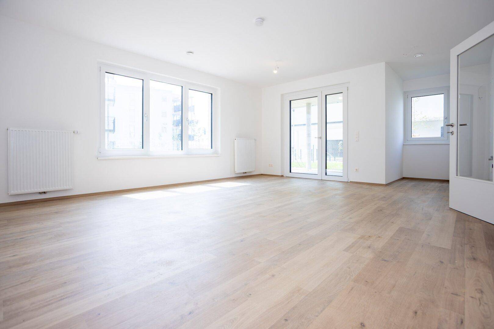 Helles, leeres Wohnzimmer mit großen Fenstern, Laminatboden und Zugang zum Außenbereich.