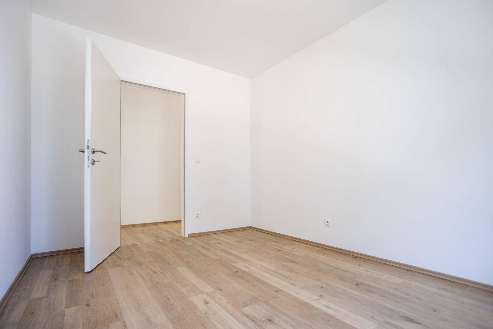Leeres Zimmer mit hellem Laminatboden und weißer Tür, bereit zur individuellen Gestaltung.