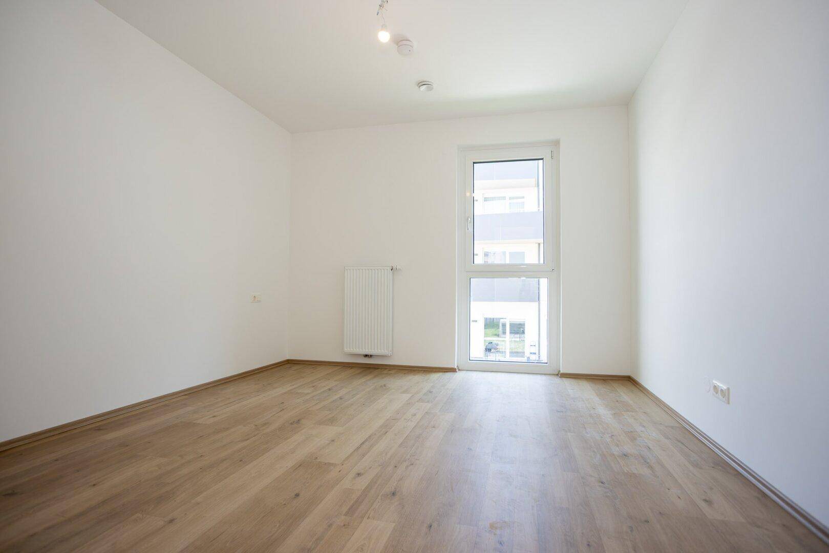 Leeres Schlafzimmer mit hellem Laminatboden und einem großen Fenster, das viel Licht hereinlässt.