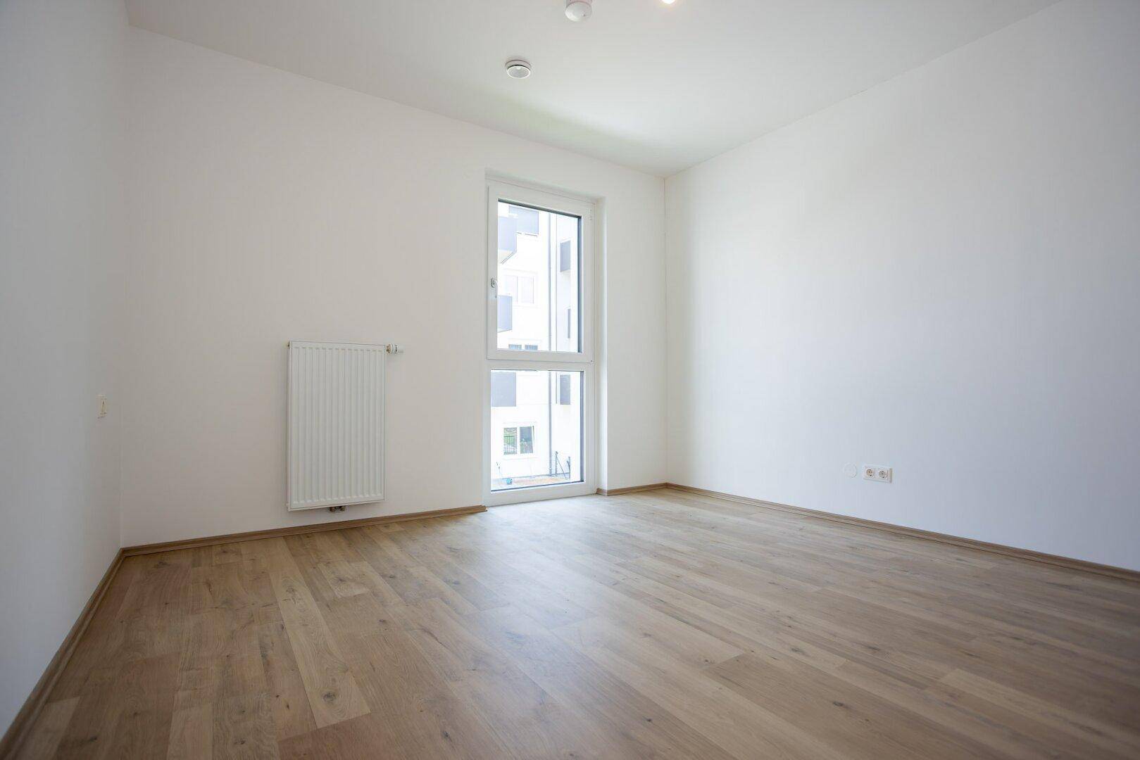 Leeres Schlafzimmer mit hellem Laminatboden und einem großen Fenster mit Blick nach draußen.