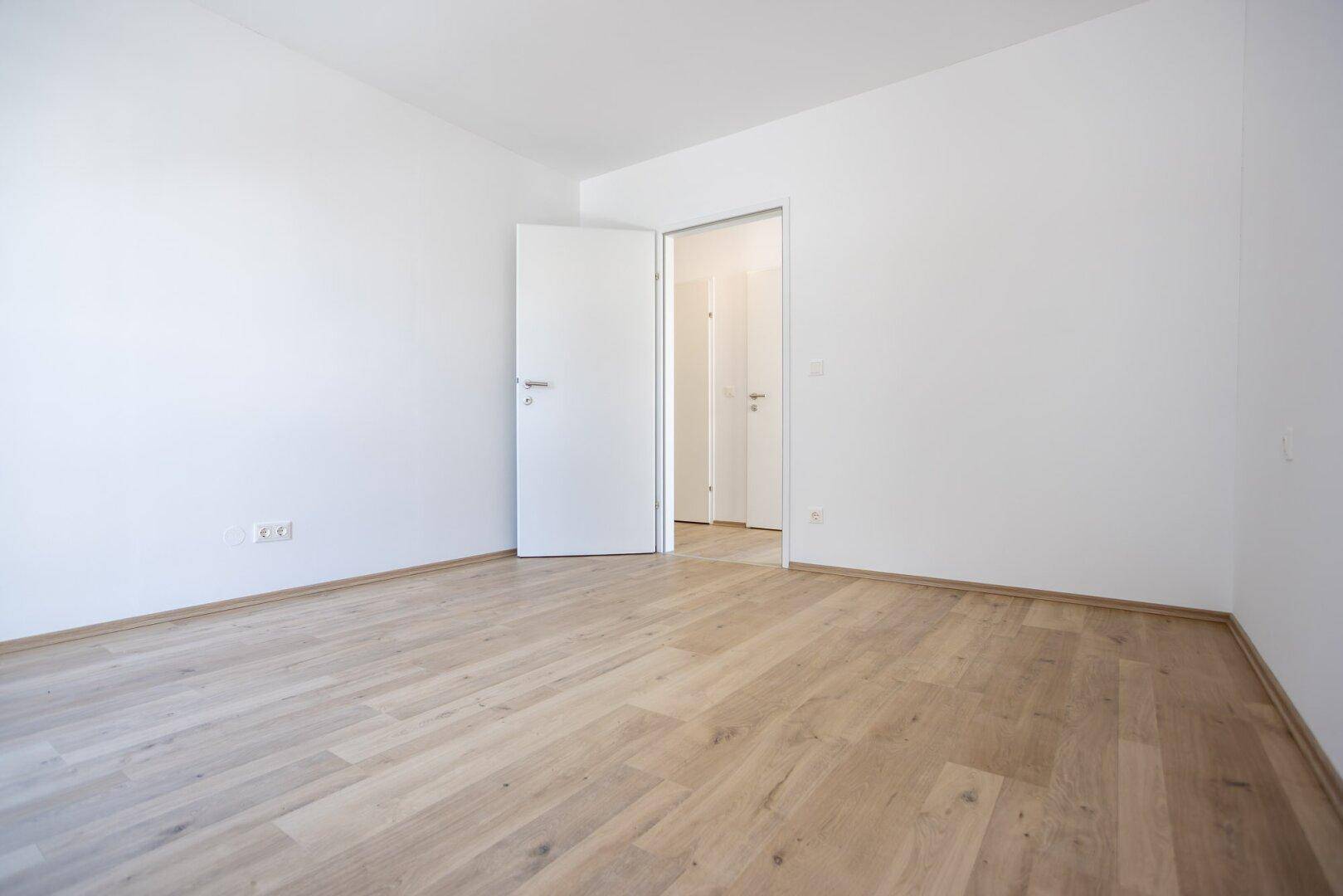Leeres Zimmer mit hellem Laminatboden und weißer Tür, bereit zur individuellen Gestaltung.