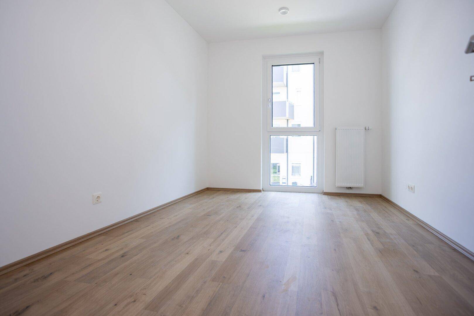 Leeres Schlafzimmer mit hellem Laminatboden und einem großen Fenster, das viel Licht hereinlässt.