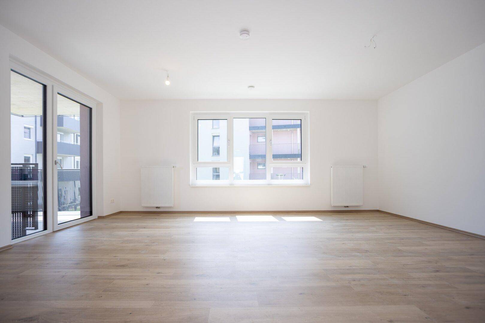 Großes, leeres Wohnzimmer mit hellem Laminatboden und weitem Blick durch die Fenster.