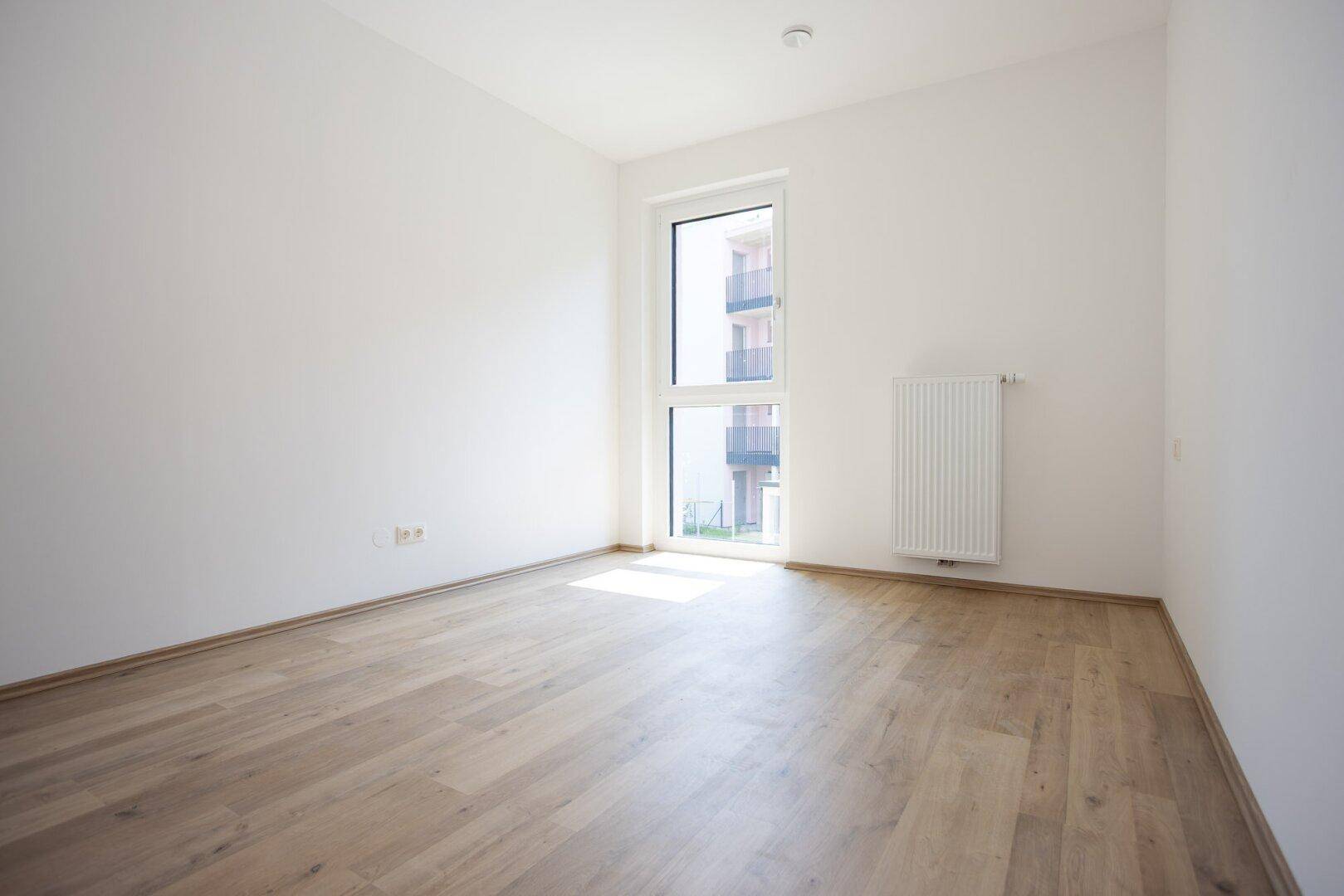 Helles Zimmer mit großem Fenster, Parkettboden und Heizkörper.