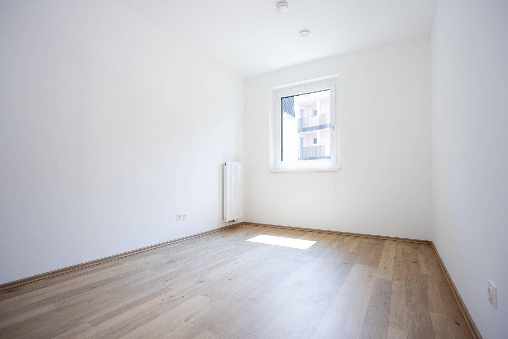 Leeres Zimmer mit Parkettboden und einem Fenster, das den Raum erhellt.