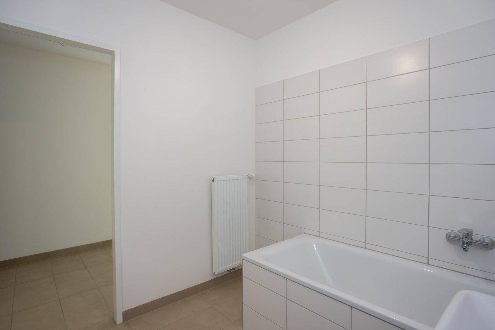 Helles Badezimmer mit Badewanne und weißen Wandfliesen, angrenzend an einen weiteren Raum.