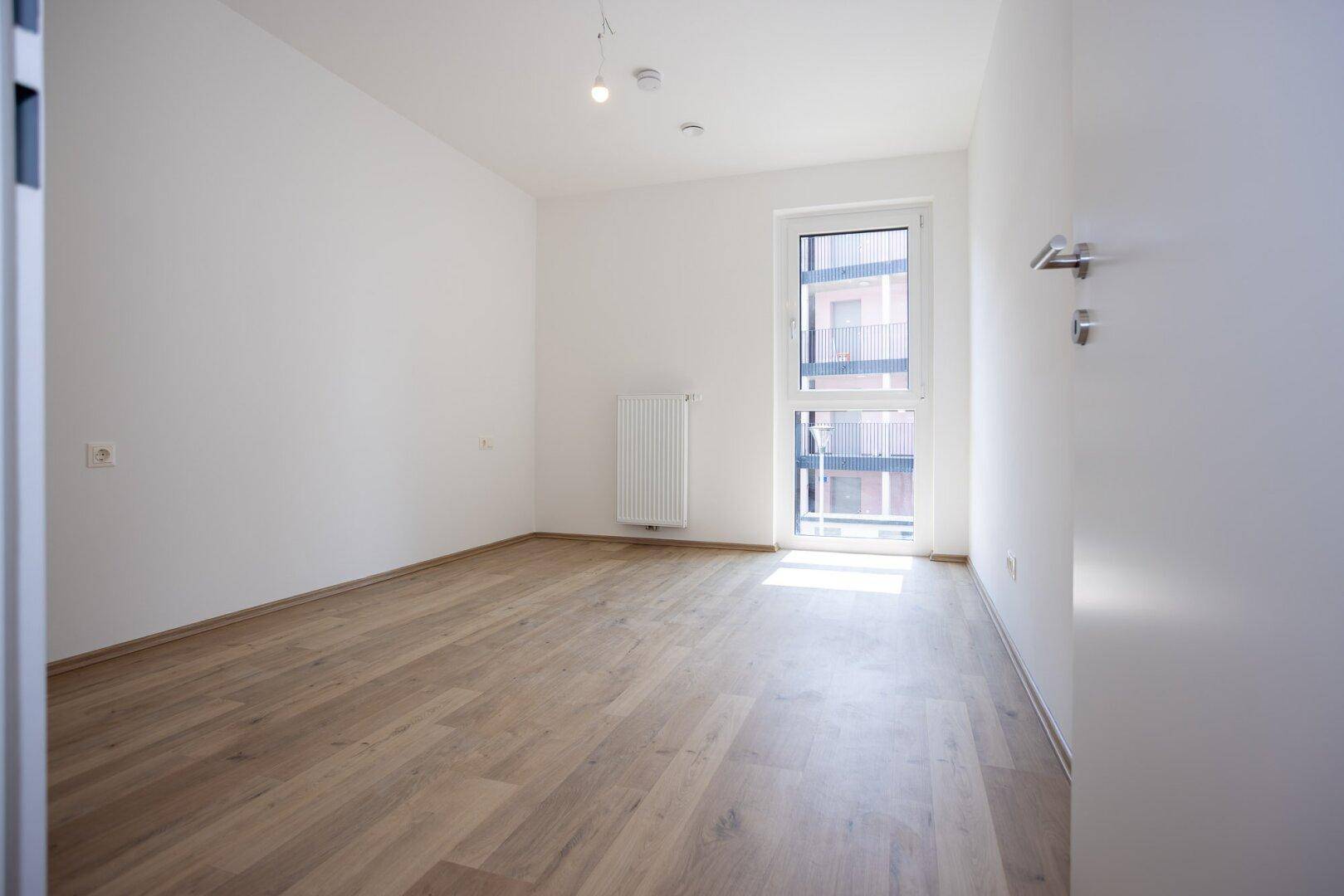 Leeres Schlafzimmer mit hellem Laminatboden, weißen Wänden und einem großen Fenster für viel Tageslicht.