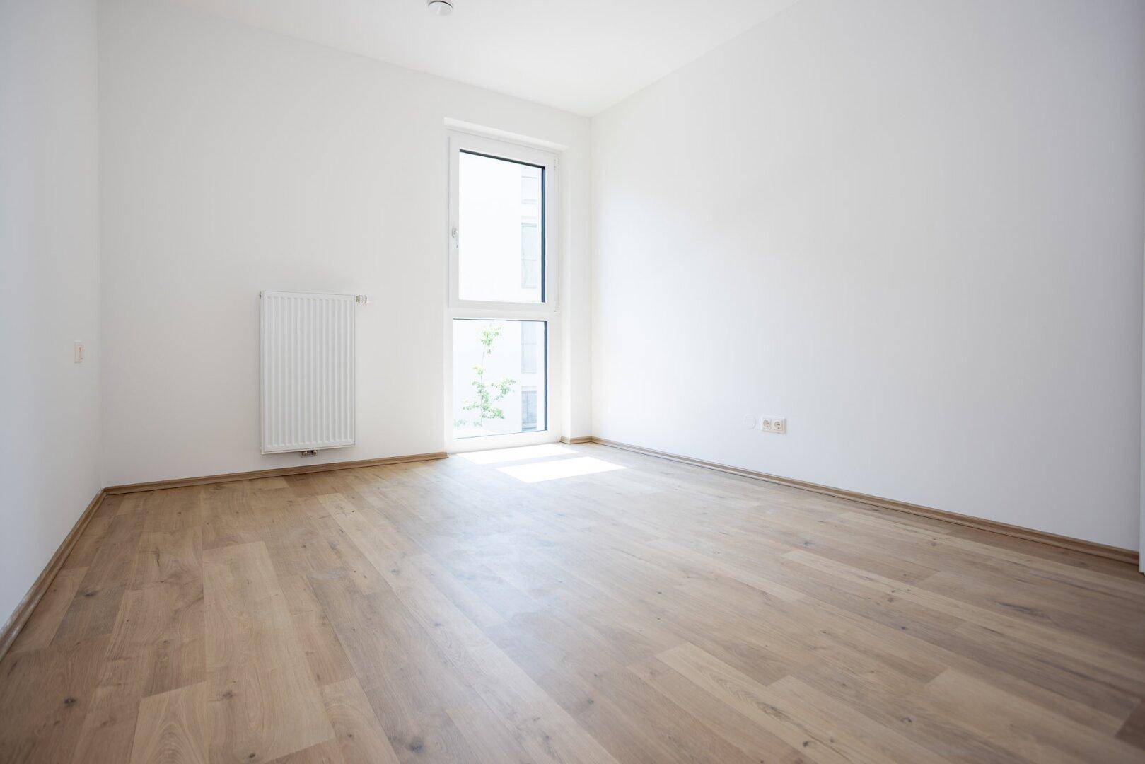 Helles, leeres Zimmer mit Laminatboden, weißen Wänden und einem hohen Fenster für viel Tageslicht.