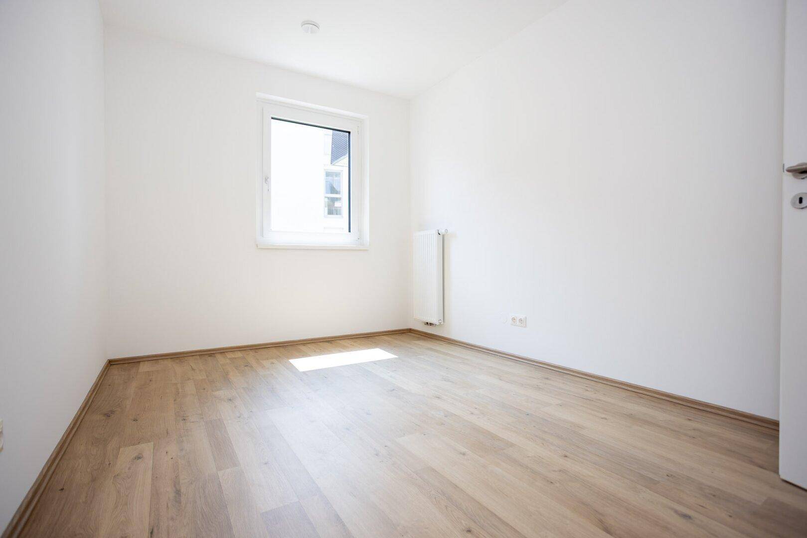 Leeres Zimmer mit hellem Laminatboden und einem Fenster, das viel natürliches Licht hereinlässt.