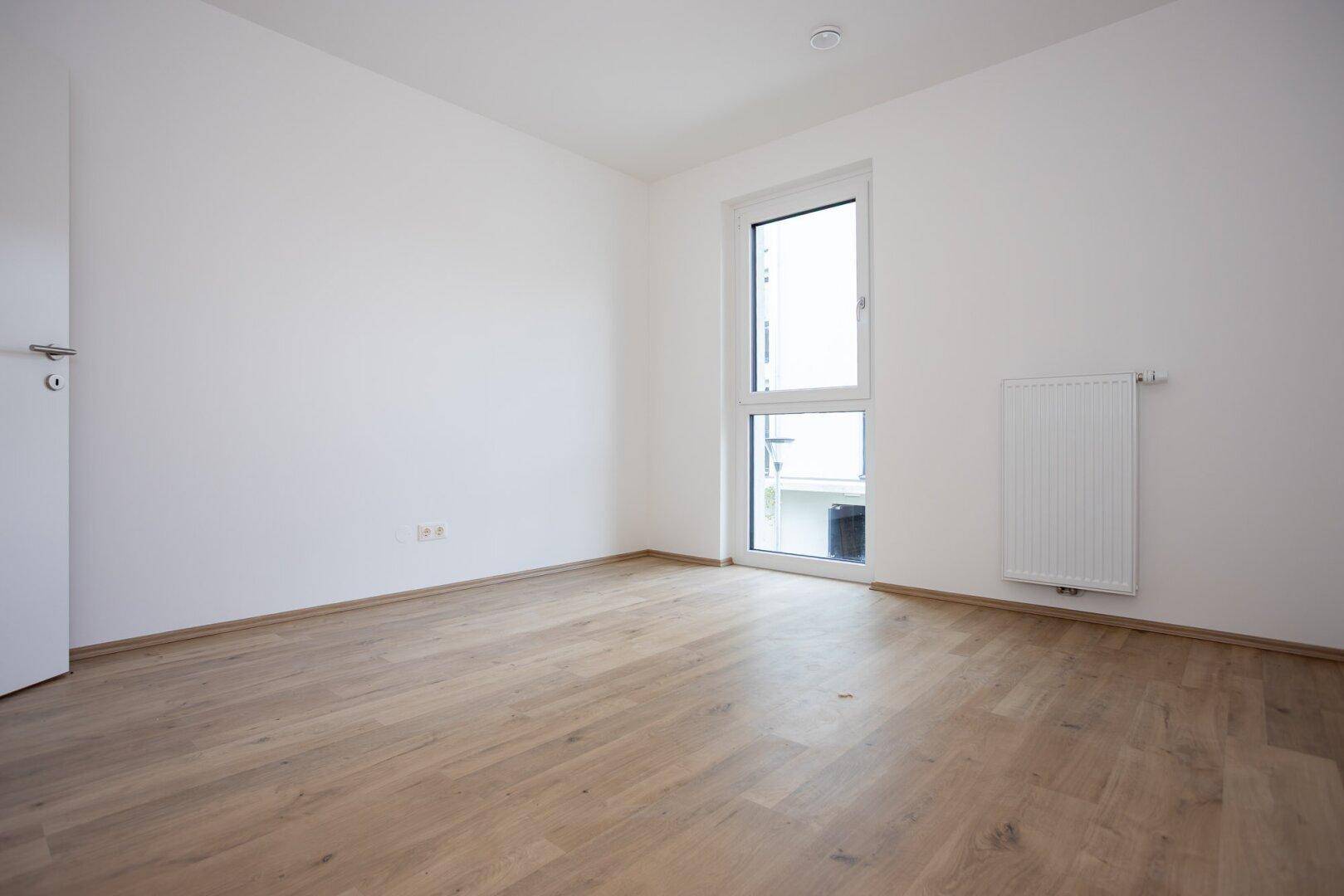 Helles Zimmer mit Laminatboden, weißen Wänden und einem Fenster mit Blick nach draußen.