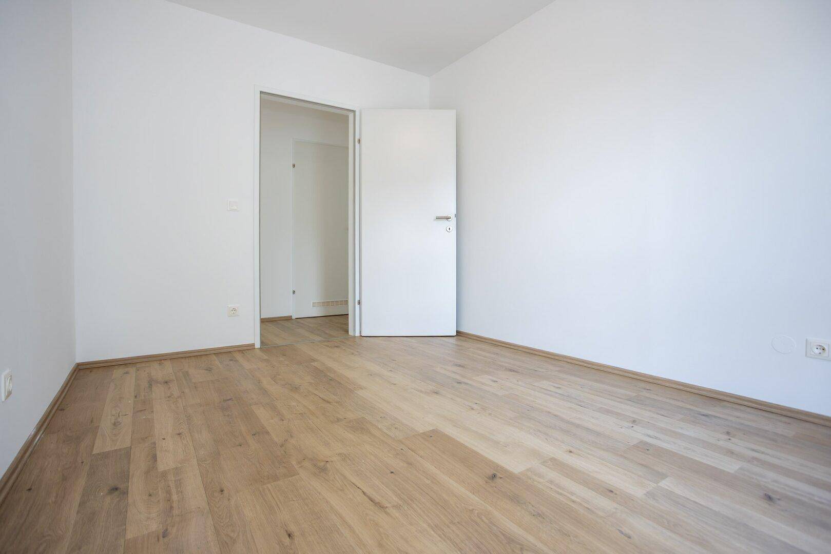 Leeres Zimmer mit Laminatboden und weiß gestrichenen Wänden, ideal als Schlaf- oder Arbeitszimmer.