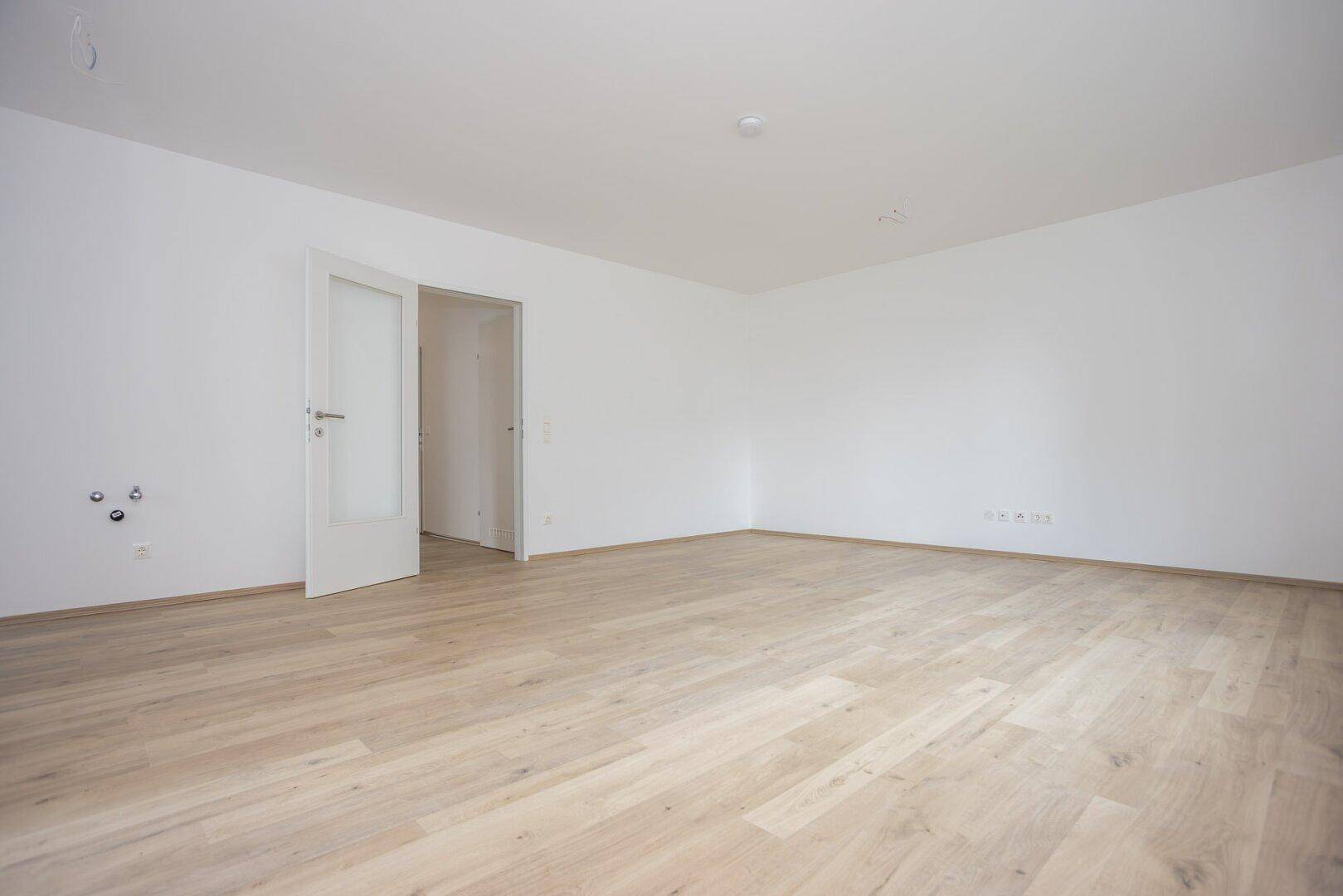 Heller, leerer Raum mit Laminatboden und Anschlüssen für eine offene Küche oder Wohnbereich.
