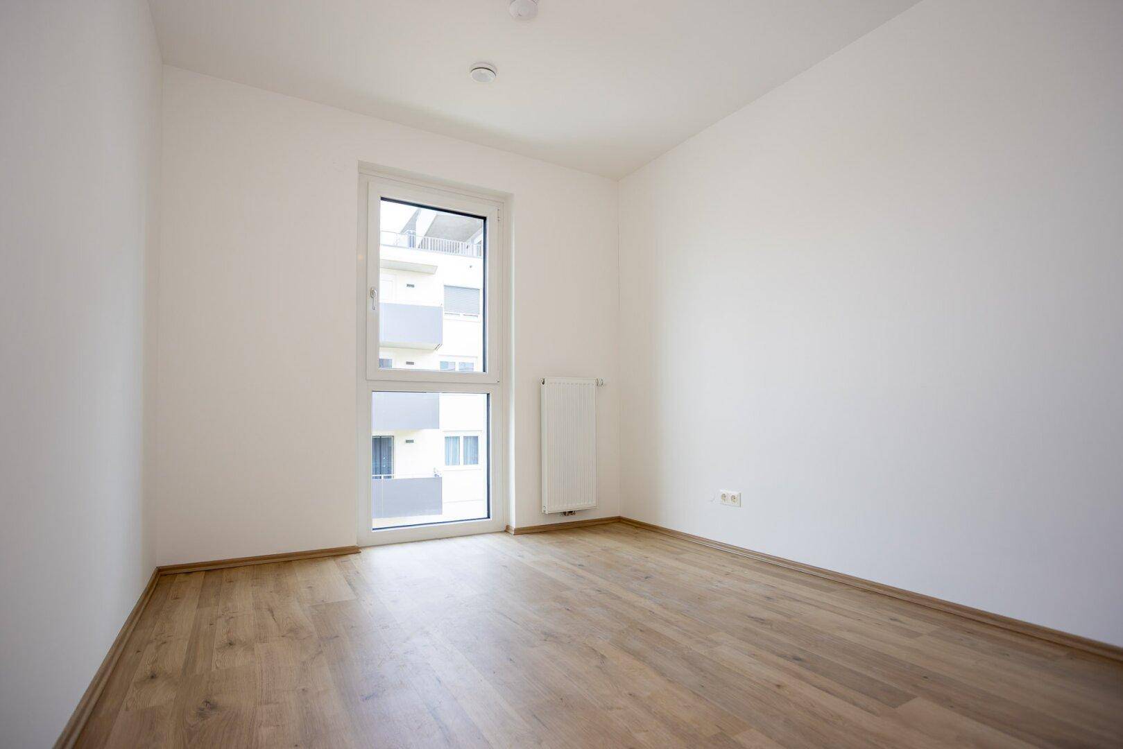 Helles Schlafzimmer mit Laminatboden, weißen Wänden und einem großen Fenster.