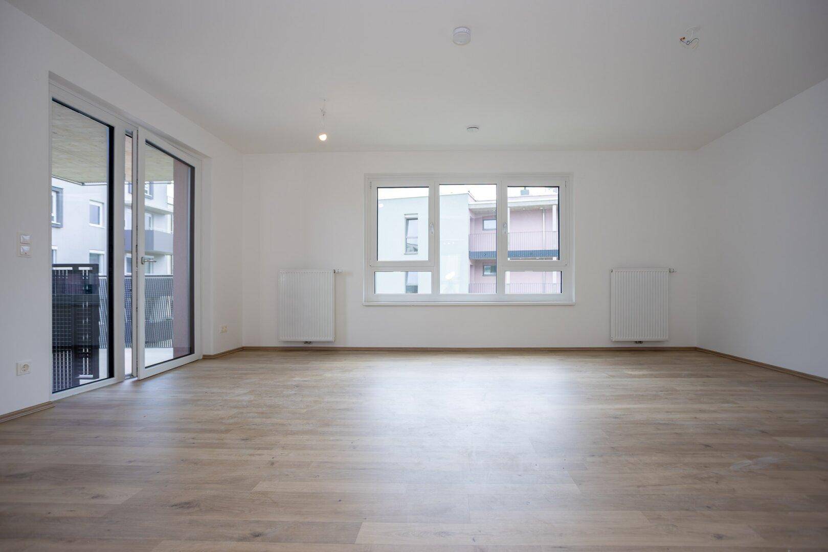 Heller Wohnbereich mit Laminatboden, großen Fenstern und direktem Zugang zum Balkon.