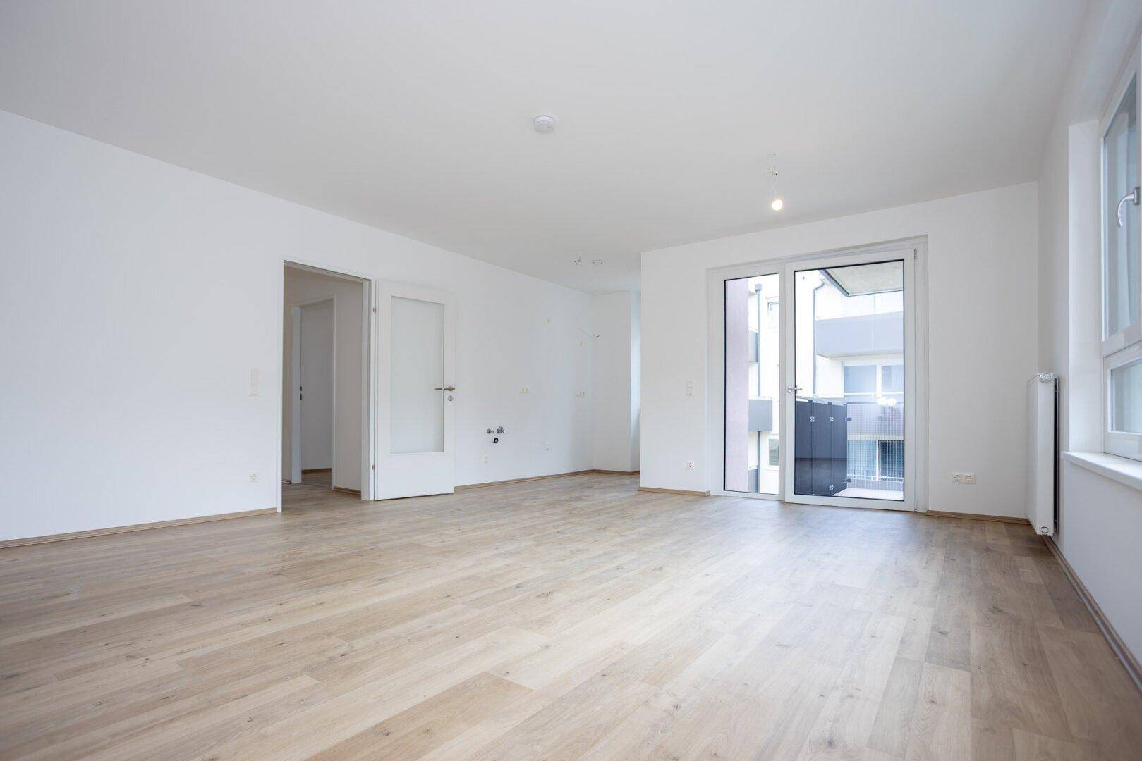 Heller Wohnbereich mit Laminatboden, weißen Wänden und direktem Zugang zum Balkon.